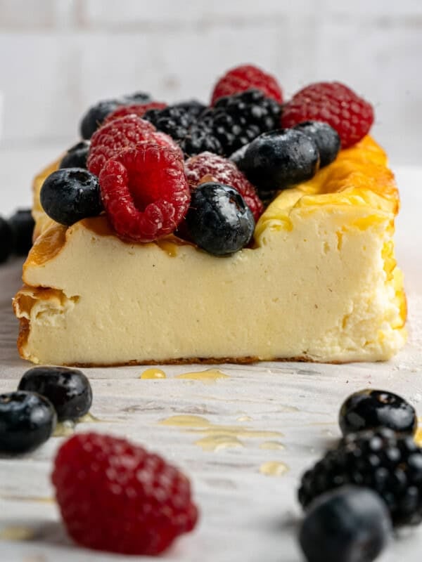 Slice of creamy cheesecake topped with fresh raspberries, blueberries, and blackberries on a white marble surface. More berries and a drizzle of honey are around the cheesecake, with a blurred white brick background.