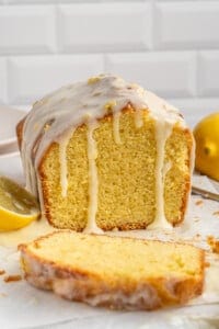Loaf of lemon drizzle cake with a thick glaze, partially sliced. A lemon wedge and knife are nearby on a white surface, with a white tiled background.