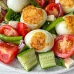 A colorful salad featuring grilled eggs, cherry tomatoes, cucumber slices, green pepper, red onion, and parsley on a white plate. The vegetables are fresh and vibrant, surrounding the eggs with a golden crust.