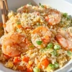 A bowl of shrimp fried rice with peas, carrots, and scrambled eggs, garnished with sesame seeds and fresh herbs. Wooden chopsticks rest on the side of the bowl. The dish is brightly lit, emphasizing its vibrant colors.