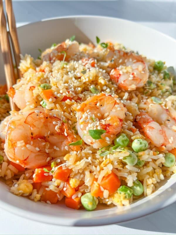 A bowl of shrimp fried rice with peas, carrots, and scrambled eggs, garnished with sesame seeds and fresh herbs. Wooden chopsticks rest on the side of the bowl. The dish is brightly lit, emphasizing its vibrant colors.