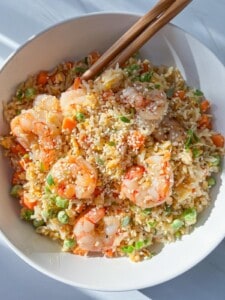 A bowl of shrimp fried rice garnished with sesame seeds and green herbs. The dish includes shrimp, rice, peas, and diced carrots, and is served with wooden chopsticks.