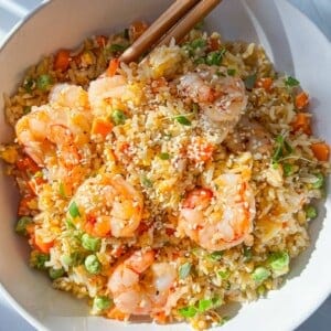 A bowl of shrimp fried rice garnished with sesame seeds and herbs. The rice is mixed with shrimp, peas, and diced carrots. Two wooden chopsticks rest on the edge of the bowl.
