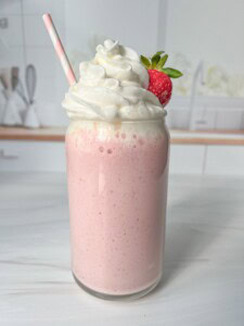 A pink strawberry milkshake in a glass jar topped with whipped cream and a fresh strawberry. A pink and white striped straw is inserted into the drink. The background features a blurred kitchen setting.