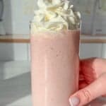 A hand holds a glass filled with a pink smoothie topped with a generous swirl of whipped cream. A red and white striped straw is placed in the drink. The smoothie is set against a light, blurred kitchen background.