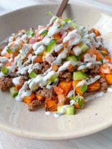 A bowl of ground beef topped with diced tomatoes, cucumbers, and drizzled with a creamy white sauce. It's garnished with fresh microgreens, served over roasted sweet potatoes on a beige plate.
