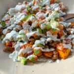 A bowl of seasoned ground beef with roasted sweet potatoes, diced tomatoes, onions, and green peppers, topped with a creamy white sauce and garnished with fresh herbs. The dish is served on a light-colored plate.