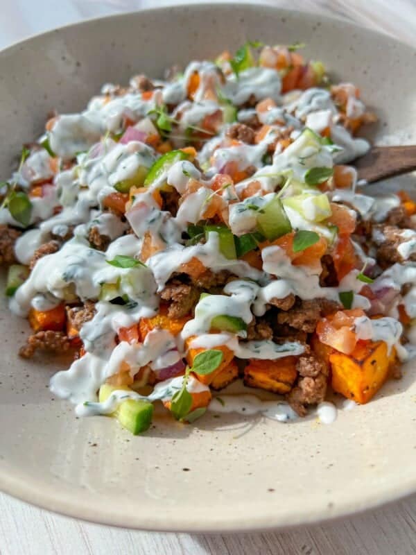A bowl of seasoned ground beef with roasted sweet potatoes, diced tomatoes, onions, and green peppers, topped with a creamy white sauce and garnished with fresh herbs. The dish is served on a light-colored plate.