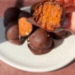 A hand holds up a chocolate-covered dessert with a bite taken, revealing a caramel or nougat-like filling. More of these treats are piled on a white plate in the background, all on a brown table surface.