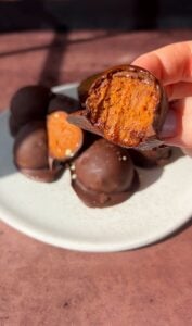 A hand holds up a chocolate-covered dessert with a bite taken, revealing a caramel or nougat-like filling. More of these treats are piled on a white plate in the background, all on a brown table surface.