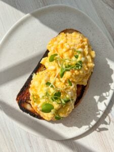 A plate with two slices of toast topped with scrambled eggs and garnished with microgreens. The toast is slightly charred, and sunlight casts shadows on the white, speckled plate and wooden table surface.