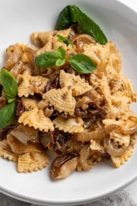 A white bowl filled with pasta topped with caramelized onions and sun-dried tomatoes. Garnished with fresh basil leaves, the dish features uniquely shaped pasta pieces with ridged edges, creating an appealing texture.