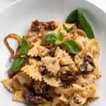 A white bowl filled with pasta topped with caramelized onions, sun-dried tomatoes, and fresh basil leaves. A small dish with grated cheese is in the background, along with basil sprigs on a light surface.