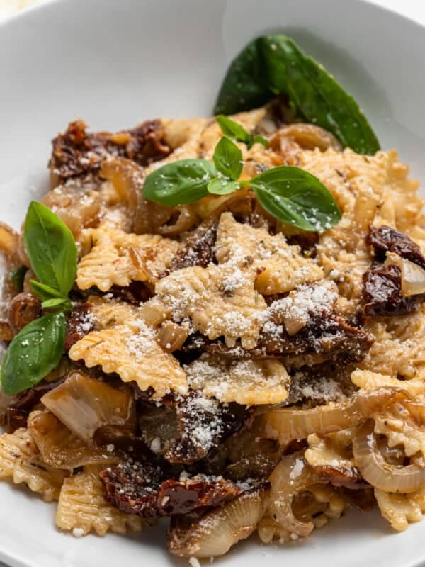 A white bowl of bow-tie pasta topped with sun-dried tomatoes, caramelized onions, grated cheese, and garnished with fresh basil leaves.