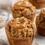 Banana muffins topped with oatmeal and peanut butter are arranged on a table. Loose oats are scattered around, and a glass bowl with oats is in the background. The muffins have a rustic, homemade appearance.