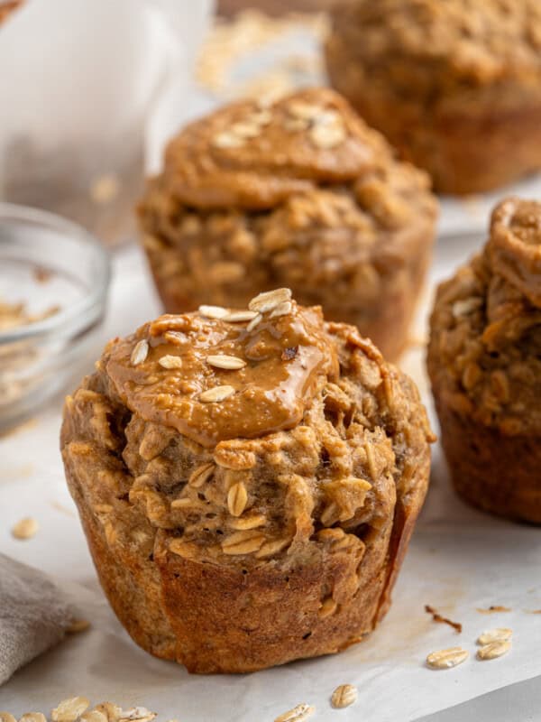 Banana muffins topped with oatmeal and peanut butter are arranged on a table. Loose oats are scattered around, and a glass bowl with oats is in the background. The muffins have a rustic, homemade appearance.