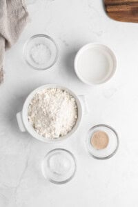 A kitchen countertop with a bowl of flour, a small bowl of salt, a cup of water, and two small bowls containing yeast and sugar. A cloth is partially visible in the top left corner.