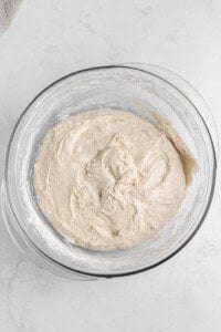 A glass bowl filled with smooth, creamy batter sits on a white marble surface. The mixture is light beige in color, with a slightly glossy texture, indicating it's well-mixed and ready for baking.