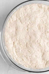 A close-up of a glass bowl containing risen, bubbly bread dough with a light, airy texture. The dough appears smooth and pale, indicating it is ready for baking. The bowl is on a white surface.