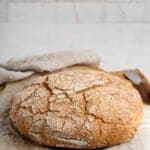 A rustic, round loaf of crusty bread sits on parchment paper atop a countertop. The bread has a cracked, golden-brown crust. A knife with a wooden handle is nearby, and a textured cloth is partially visible in the background.