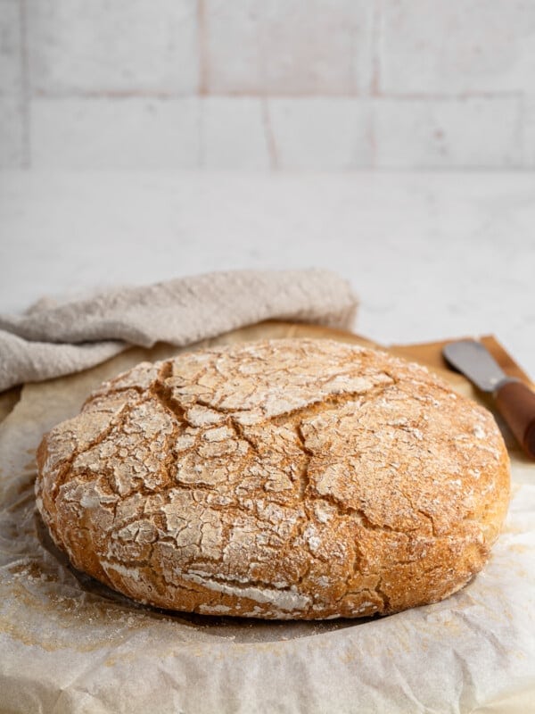 A rustic, round loaf of crusty bread sits on parchment paper atop a countertop. The bread has a cracked, golden-brown crust. A knife with a wooden handle is nearby, and a textured cloth is partially visible in the background.