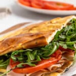 A freshly made sandwich on a wooden board, filled with sliced turkey, fresh arugula, and tomato slices. The sandwich is on ciabatta bread, slightly toasted. A white plate with additional tomato slices is blurred in the background.