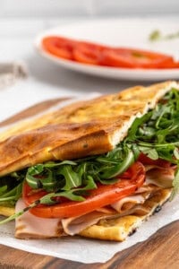 A freshly made sandwich on a wooden board, filled with sliced turkey, fresh arugula, and tomato slices. The sandwich is on ciabatta bread, slightly toasted. A white plate with additional tomato slices is blurred in the background.