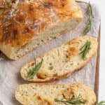 Freshly baked rosemary bread on a wooden board with parchment paper. One whole round loaf and two slices are visible, garnished with sprigs of rosemary. The crust is golden and the crumb looks soft and airy.