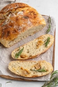 Freshly baked rosemary bread on a wooden board with parchment paper. One whole round loaf and two slices are visible, garnished with sprigs of rosemary. The crust is golden and the crumb looks soft and airy.