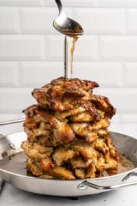 A stack of grilled chicken pieces skewered on a spit in a pan. A spoon drizzles sauce over the top, enhancing the golden-brown, juicy appearance of the chicken. The background features a white, tiled wall design.