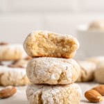 A stack of three powdered almond cookies on a wooden surface. The top cookie has a bite taken out, revealing a soft interior. Whole almonds are scattered around, with more cookies blurred in the background.