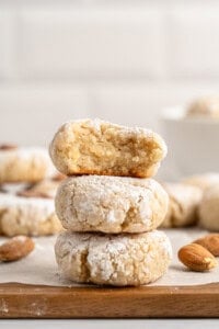 A stack of three powdered almond cookies on a wooden surface. The top cookie has a bite taken out, revealing a soft interior. Whole almonds are scattered around, with more cookies blurred in the background.
