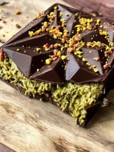 A close-up of a piece of chocolate with a unique, angular top design. It is topped with small bits of crushed nuts, and the center reveals a green, textured filling on a wooden surface.