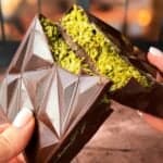 Two hands hold a triangular chocolate bar filled with a textured green filling. The chocolate has a geometric design, and the filling appears to be a finely shredded, vibrant substance, possibly pistachio. The background is blurred and warm-toned.