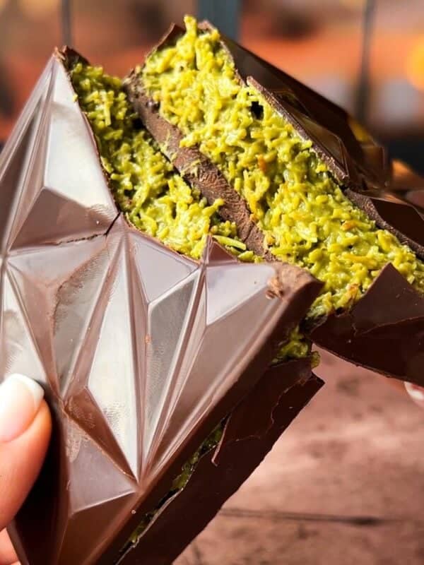 Two hands hold a triangular chocolate bar filled with a textured green filling. The chocolate has a geometric design, and the filling appears to be a finely shredded, vibrant substance, possibly pistachio. The background is blurred and warm-toned.