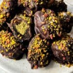 A plate of chocolate-covered dates sprinkled with crushed nuts and colorful toppings. One of the dates is cut open, revealing a green filling. The plate is on a light wooden surface.