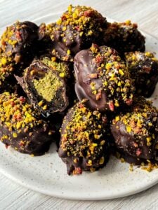 A plate of chocolate-covered dates sprinkled with crushed nuts and colorful toppings. One of the dates is cut open, revealing a green filling. The plate is on a light wooden surface.