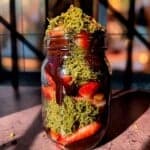 A jar filled with fresh sliced strawberries, melted chocolate, and shredded green topping, possibly pistachios or matcha, sits on a surface with a blurred, backlit background.