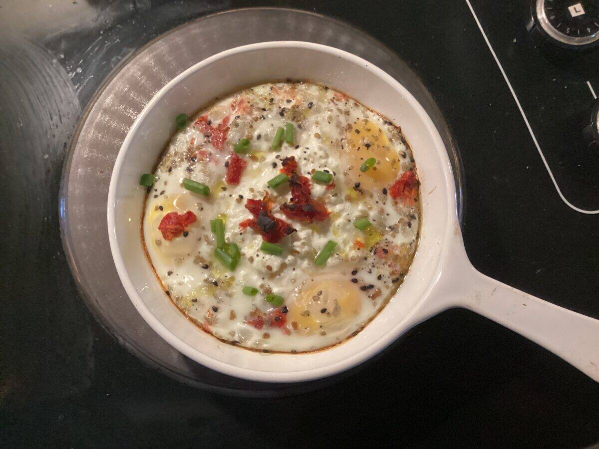 A white pan with baked eggs, topped with chopped green onions, sun-dried tomatoes, black pepper, and seasonings, sits on a black stovetop.