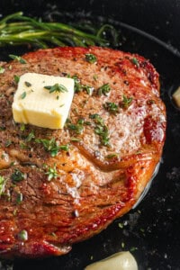 Close-up of a sizzling steak topped with a pat of melting butter and garnished with fresh thyme. The steak is seared with a caramelized crust, and sprigs of rosemary are visible in the background.