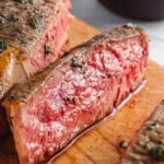 Close-up of juicy, medium-rare steak slices on a wooden cutting board, garnished with herbs and pepper. A blurred bowl with herbs is in the background.
