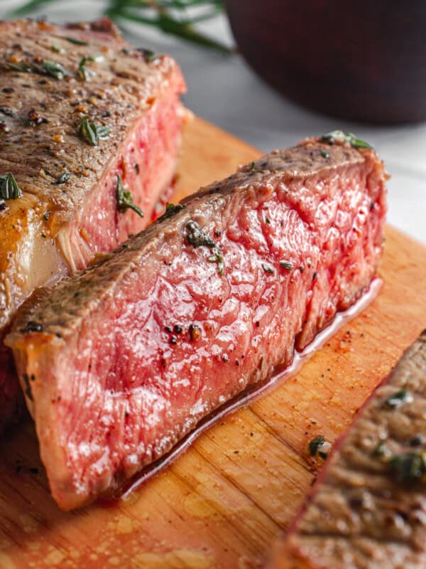 Close-up of juicy, medium-rare steak slices on a wooden cutting board, garnished with herbs and pepper. A blurred bowl with herbs is in the background.