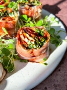 Close-up of a plate of prosciutto-wrapped rolls filled with greens, sun-dried tomatoes, and cheese, garnished with microgreens. The dish is presented on a speckled white plate, with light highlighting the vibrant colors.