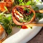 Close-up of Italian sushi recipe wrapped in prosciutto, filled with greens, and drizzled with a dark sauce on a white plate. They're accompanied by small leafy greens, with chopsticks on the side. The setting is warmly lit.
