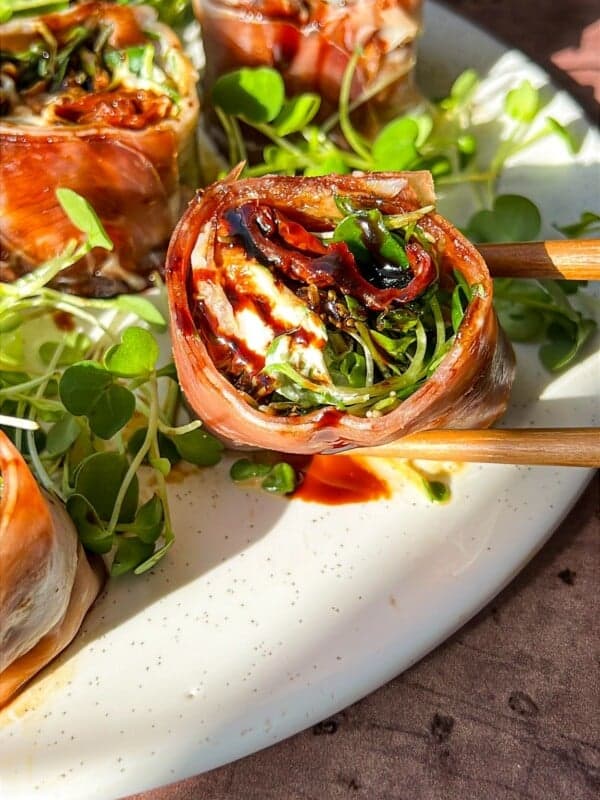 Close-up of Italian sushi recipe wrapped in prosciutto, filled with greens, and drizzled with a dark sauce on a white plate. They're accompanied by small leafy greens, with chopsticks on the side. The setting is warmly lit.