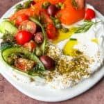 A plate featuring creamy labneh topped with smoked salmon, cherry tomatoes, cucumber ribbons, olives, capers, and fresh dill, drizzled with olive oil and sprinkled with za'atar.