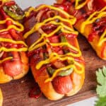 Three grilled Mexican Hot Dogs in buns topped with ketchup, mustard, grilled onions, and green bell peppers, arranged on a wooden serving board. A sprig of parsley is visible in the corner.