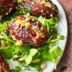 Close-up of stuffed dates on a plate, filled with a crumbly mixture. They are placed on a bed of fresh greens and sprinkled with pine nuts, on a rustic brown background.