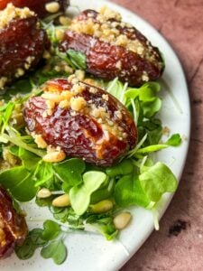 Close-up of stuffed dates on a plate, filled with a crumbly mixture. They are placed on a bed of fresh greens and sprinkled with pine nuts, on a rustic brown background.
