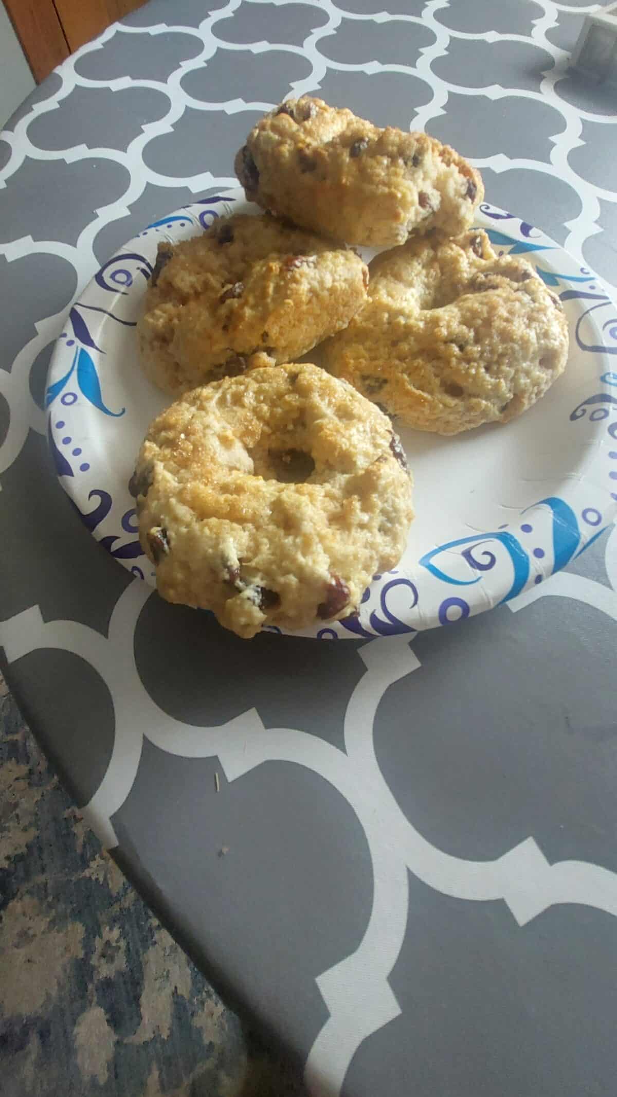 A paper plate with four homemade, rustic-looking scones or biscuits sits on a patterned grey and white table. The scones appear golden brown with visible bits of fruit or nuts.